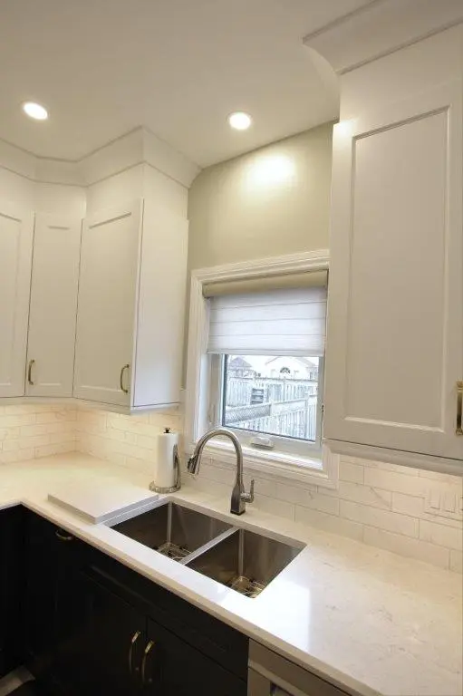 Modern kitchen corner with white quartz countertop, undermount double stainless steel sink, silver faucet, white cabinetry above and dark cabinetry below, window with white blind, and soft ceiling lighting.