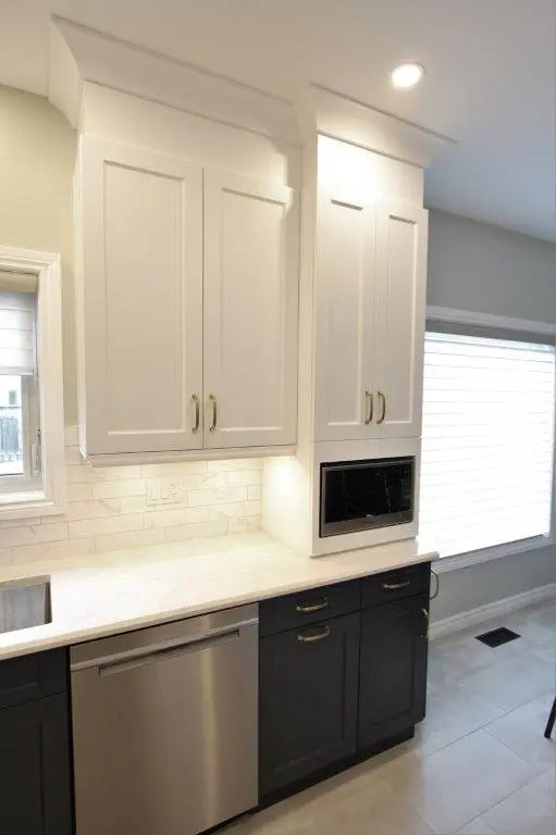 Modern kitchen corner with white upper cabinets and dark lower cabinets, stainless steel dishwasher, built-in microwave, white subway tile backsplash, and a window with a white blind illuminating the area.