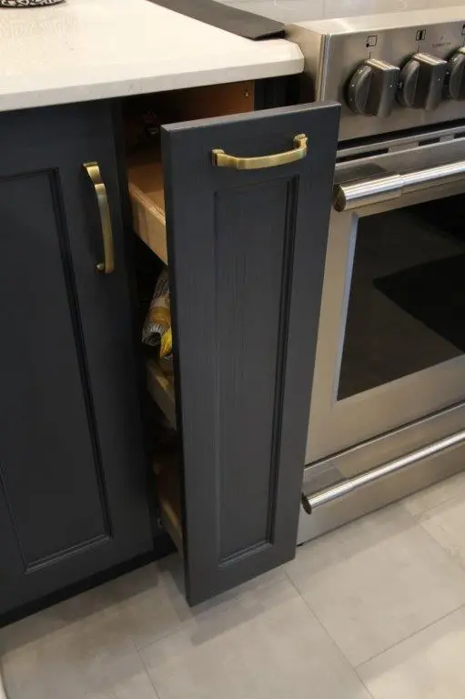 Narrow kitchen cabinet drawer with dark gray door and brass handle, partially open showing storage, next to a stainless steel oven with control knobs and handle.