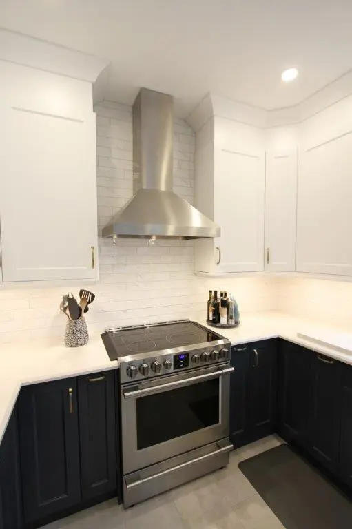 Modern kitchen corner with stainless steel electric stove and range hood, white upper cabinets, dark lower cabinets, white tiled backsplash, countertop utensils, and bottles.