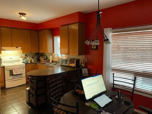 A before picture of a Kitchen and dining area with red walls, wooden cabinets, a white stove, a countertop with chairs, and a round table with laptops and office supplies near a window with blinds.