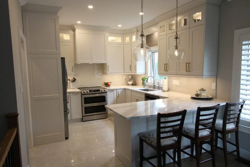 Contemporary kitchen with white cabinetry, quartz countertops, stainless steel appliances, three wooden bar stools with patterned cushions, two pendant lights with glass shades, and a window with white shutters.