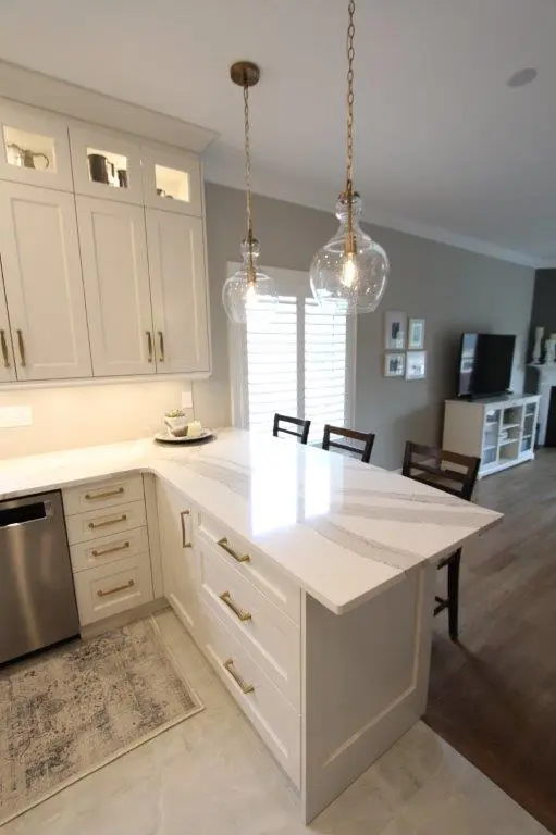 Bright kitchen corner with white cabinetry, quartz countertop island with gold hardware, two hanging glass pendant lights, a dishwasher, and a small decorative plant on a tray, adjacent to a living room with a TV and chairs.