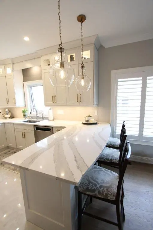 Modern kitchen with a white quartz countertop island, three dark wooden bar stools with patterned cushions, pendant lights with glass globes hanging above, and white cabinetry with gold handles under soft lighting.