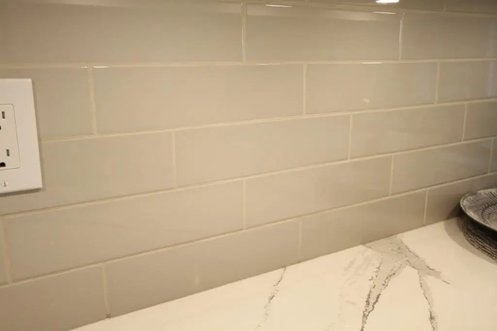 Close-up of a kitchen backsplash with beige rectangular subway tiles, a white electrical outlet on the left, and a quartz countertop with gray veining on the bottom right corner.