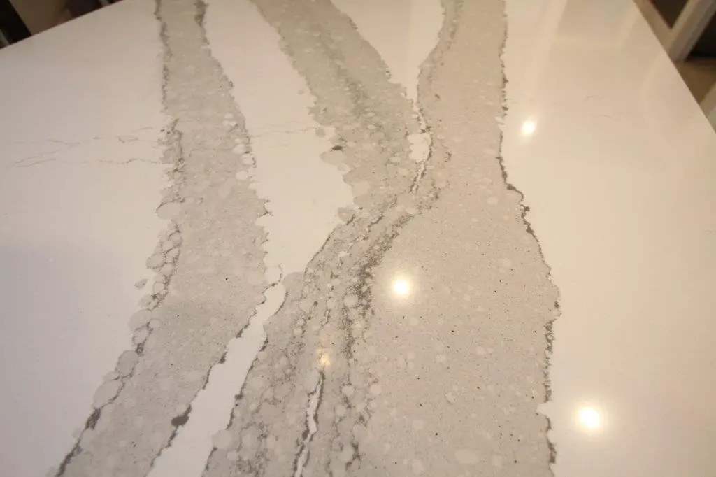 Close-up of a smooth white surface with three wide, irregular, light gray vein-like patterns running vertically, resembling quartz countertop, with a few reflections of overhead lights.
