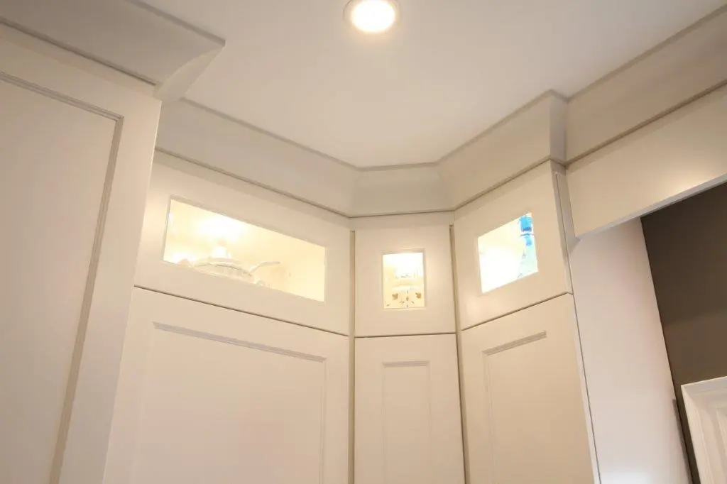 Ceiling corner with recessed lighting and upper cabinet doors featuring small illuminated display windows showing decorative objects inside.