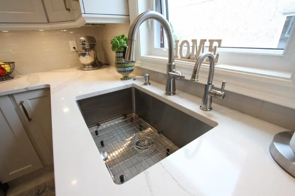 Modern kitchen sink with dual stainless steel faucets, white quartz countertop, a small decorative plant, and wooden letters spelling 'HOME' on the windowsill.