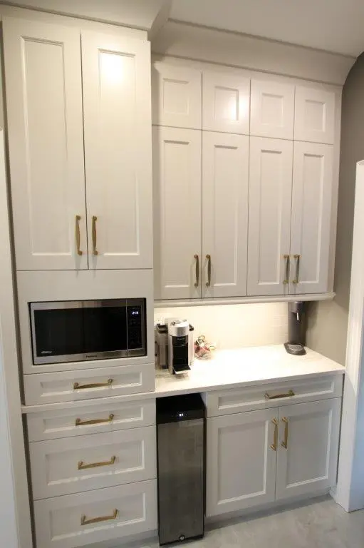 White kitchen cabinetry with brass handles, featuring an integrated microwave, a coffee machine on the countertop, and a stainless steel trash bin below the counter within a modern kitchen setup.