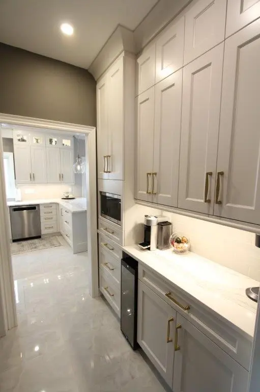 Modern kitchen interior with white cabinetry featuring gold handles, quartz countertops, built-in appliances, a coffee maker, and a glass bowl of fruit on the counter, with a view into an adjacent kitchen area with additional cabinetry and stainless steel dishwasher.