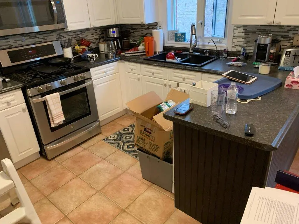 A before picture of a kitchen with stainless steel stove and white cabinets, a countertop with various items including a cardboard box, water bottle, glasses, and a tablet, tiled floor with a small patterned rug near the stove, and a window above the sink.