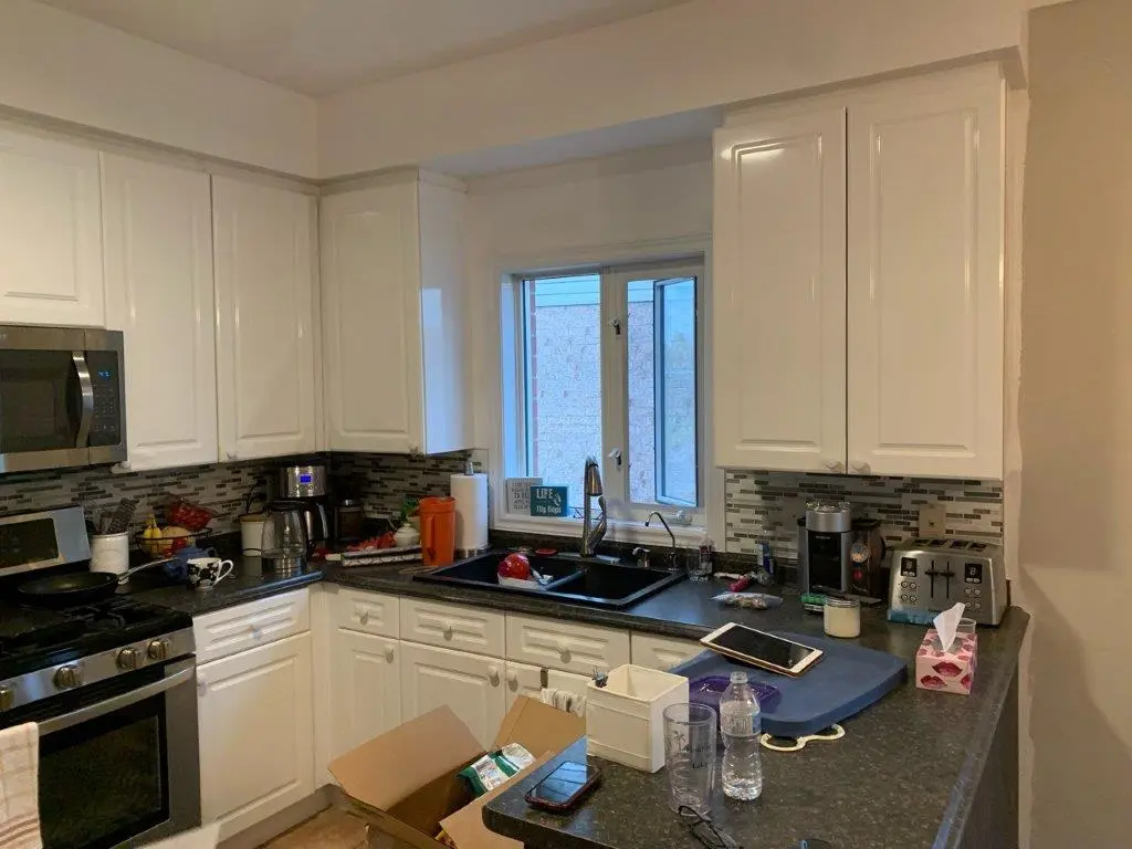A before picture of a kitchen with white cabinets, black countertops, a double black sink under a window, various kitchen appliances including a toaster, coffee maker, microwave, and stove, a cardboard box on the counter, a water bottle, some utensils, and a tablet on a blue mat.
