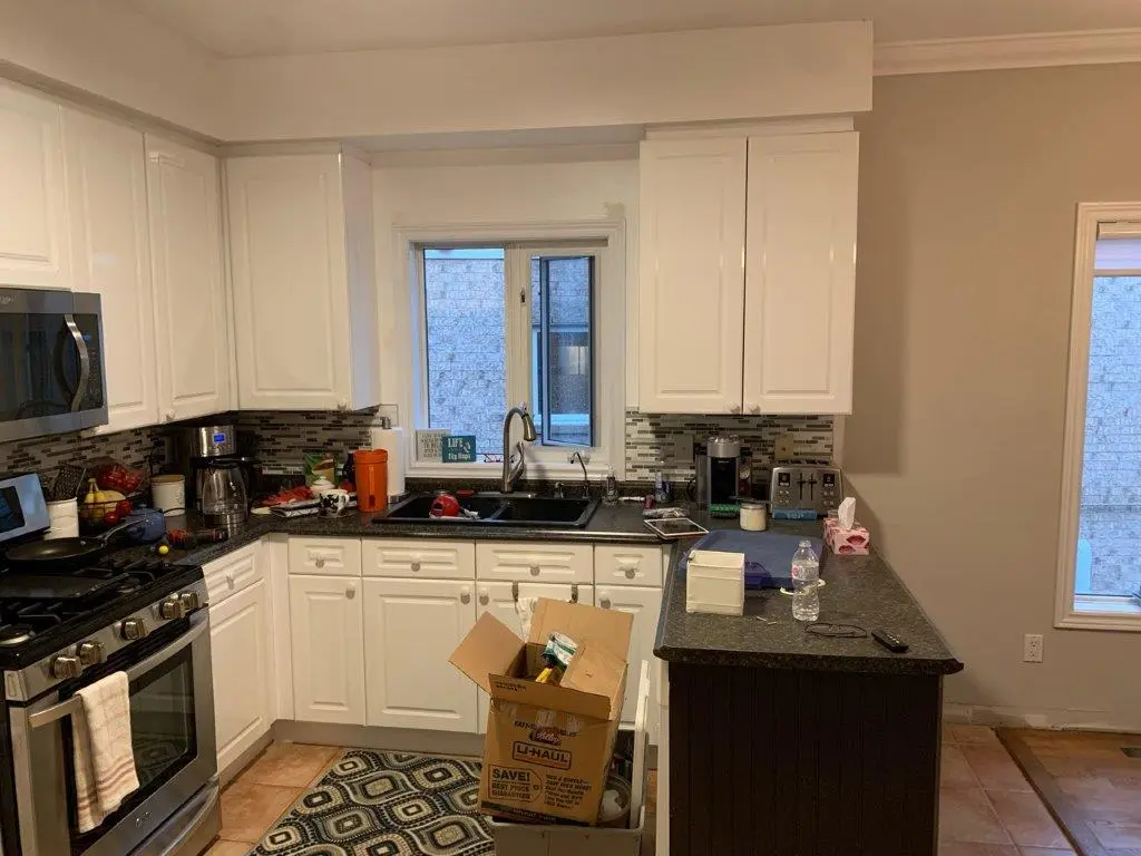 A before picture of a kitchen with white cabinets, black countertops, and a window above the sink; includes stainless steel stove and microwave, coffee maker, toaster, and various kitchen items; a patterned rug is on the floor, and an open U-Haul cardboard box is on the floor in front of the counter.