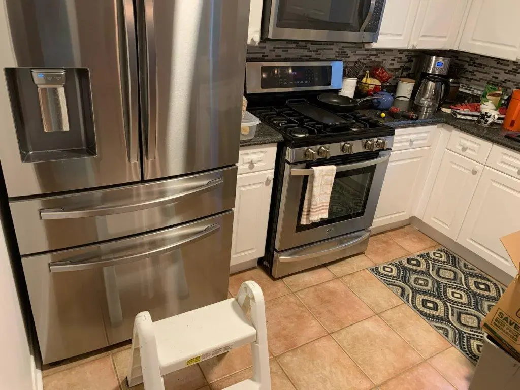 A before picture of a kitchen with stainless steel refrigerator and stove, white cabinets, tiled floor, countertop with various kitchen items, and a small step stool in front of the refrigerator.