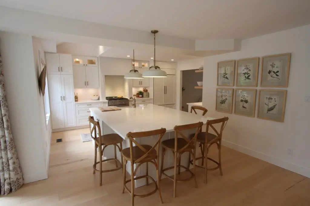 A modern kitchen featuring a large white island with wooden chairs, pendant lights above, and white cabinetry with stainless steel appliances. Botanical artwork is displayed on the wall.