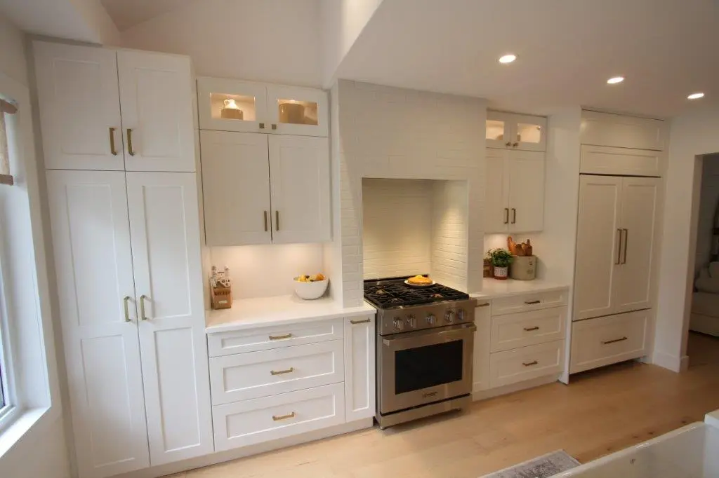 A modern kitchen featuring white cabinetry, a stainless steel oven with a gas range, and built-in appliances, all under recessed lighting on a light wood floor.