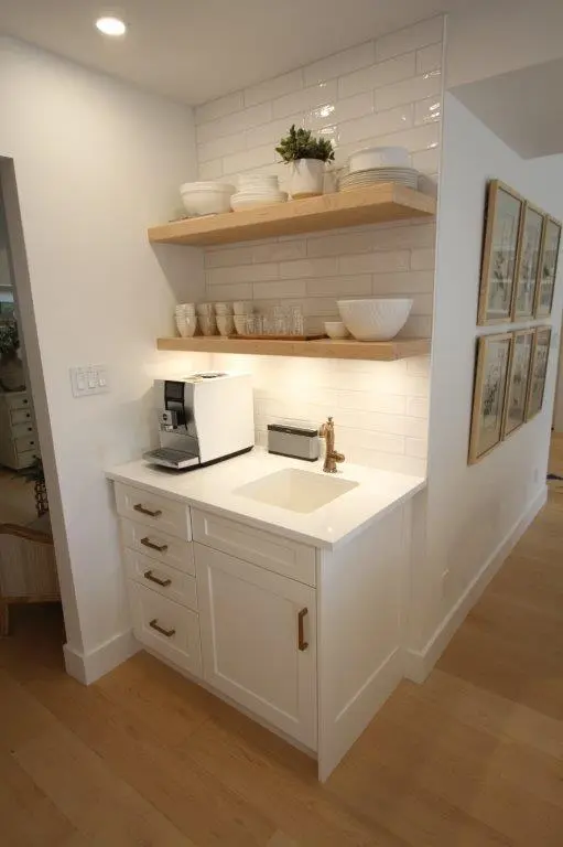 A small white kitchen nook featuring a countertop with a built-in sink, a modern coffee machine, and floating wooden shelves displaying white dishes and a small potted plant. The backsplash is composed of white subway tiles, and there are framed pictures hung on the adjacent wall. The flooring is light wood, complementing the clean and minimalist design.