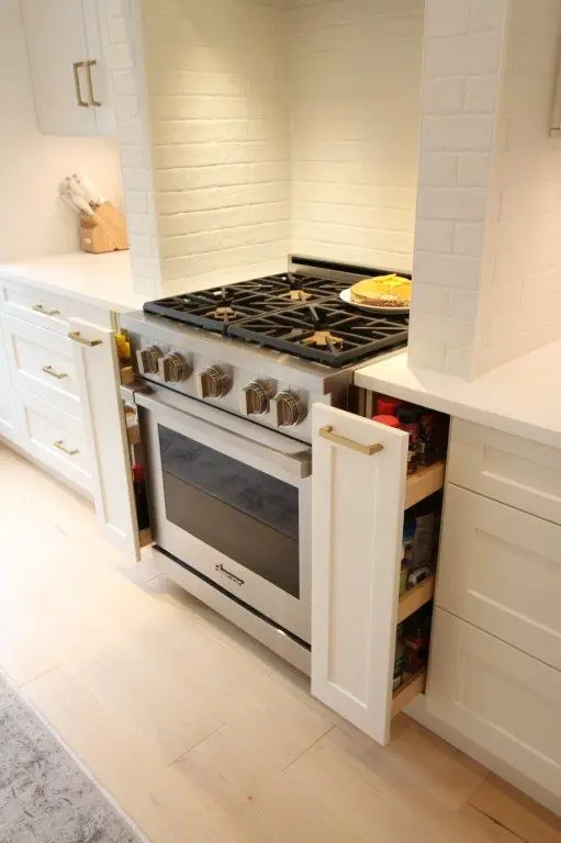 A modern kitchen with a stainless steel gas stove and oven. The stove is flanked by two pull-out spice racks integrated into the white cabinetry with gold handles. The backsplash is made of white brick tiles, and a pie is placed on the counter behind the stove.