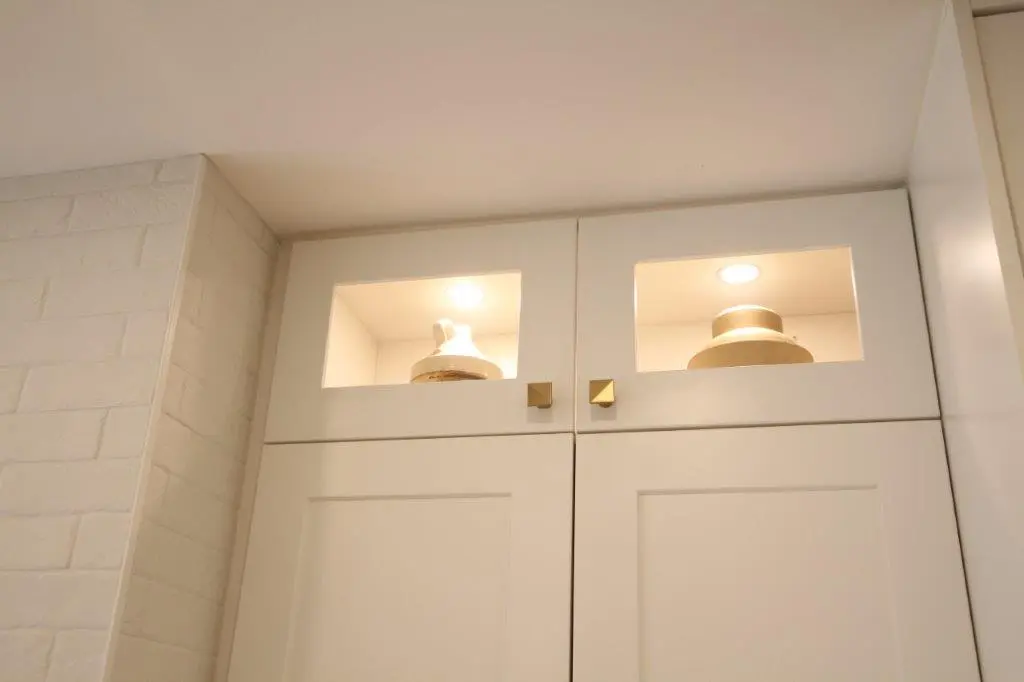 White kitchen cabinets with glass panel doors at the top displaying decorative pottery, lit by small overhead lights, next to a brick wall.
