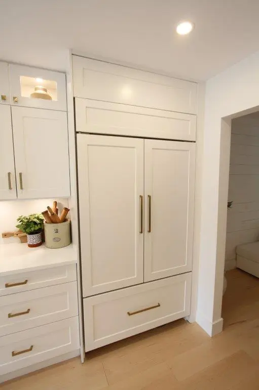 Built-in white kitchen fridge and cabinetry with paneled doors and brass handles, a countertop with potted herbs and wooden utensils, and light wood flooring under recessed ceiling light