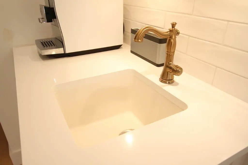 White countertop with an under-mounted square sink, a brass faucet, a white coffee machine, and a silver speaker against a white tiled backsplash.