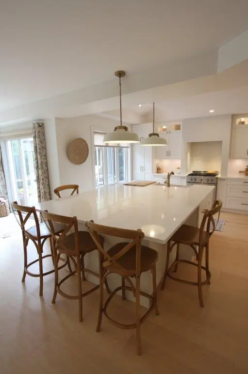Bright modern kitchen with a large white island countertop surrounded by six wooden bar stools with cross-back design, pendant lights hanging above, light wood flooring, and white cabinetry with a stove in the background.