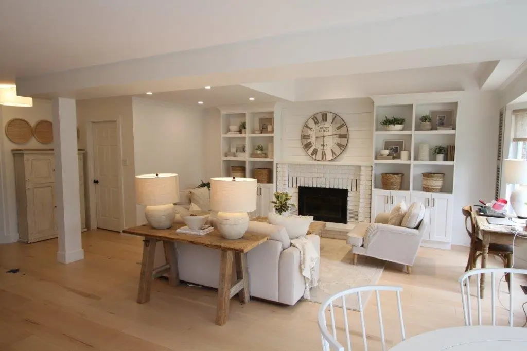 Bright and cozy living room with light wooden floors, a beige sofa and armchair, a wooden console table with two lamps behind the sofa, built-in white shelving around a white brick fireplace with a large round clock above it, and a round white dining table in the foreground.