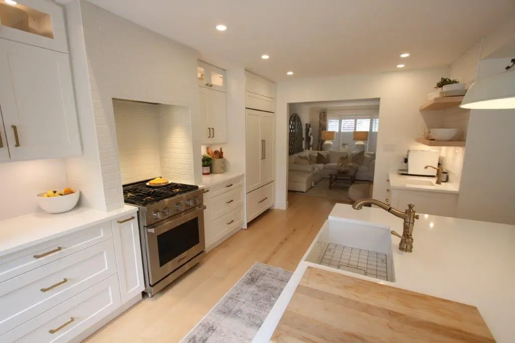 Modern kitchen with white cabinetry, stainless steel stove, light wood flooring, brass faucet, and a view into a cozy living room with a sectional sofa and warm lighting.