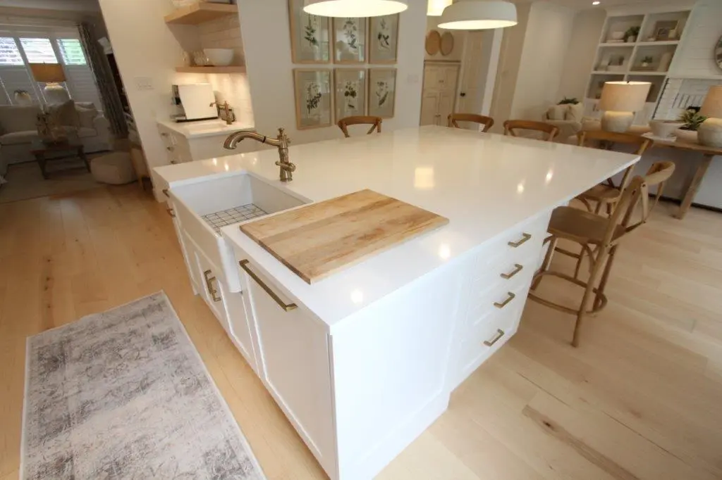 A modern kitchen with a large white island featuring a built-in sink, brass faucet, and a wooden cutting board. The kitchen has light wooden floors, white cabinetry, and warm lighting fixtures above. There are chairs around the island and additional seating at a nearby table.