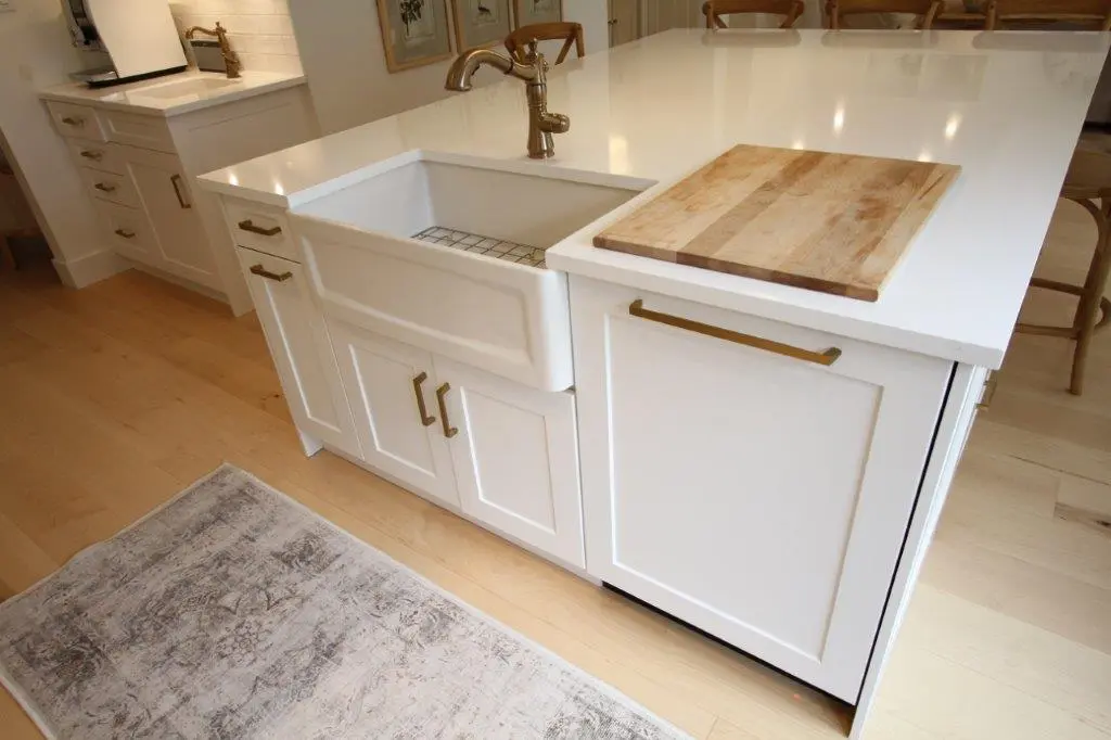 A modern kitchen island with a farmhouse sink, brass faucet, and cabinetry with brass handles, featuring a wooden cutting board on the countertop and a beige rug on the light wooden floor.