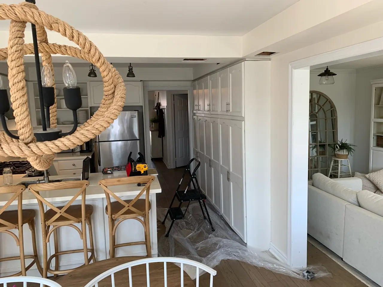 A before renovation kitchen and living room area with a light fixture made of rope, wooden stools at a kitchen island, and a set of white cabinets against one wall. A step ladder and tools are on the floor, indicating ongoing remodeling or maintenance work. The adjacent living room features a white sofa and a large rustic mirror.