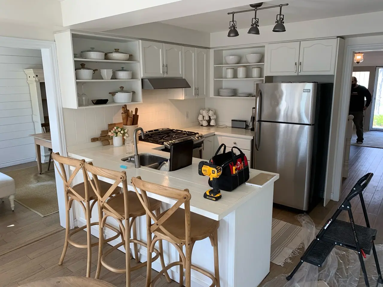 A before renovation kitchen with white cabinetry, open shelves, and a stainless steel refrigerator. The counter has a small sink. Three wooden bar stools are placed by the counter.