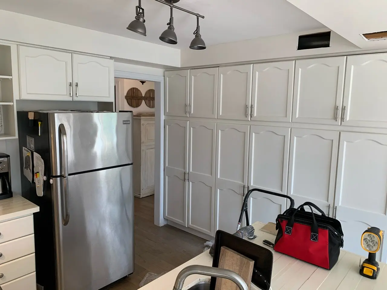 A before renovation kitchen with white cabinets, a stainless steel refrigerator, and a table with a red and black tool bag, step ladder, and flashlight on it.