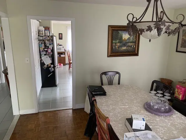 A before renovation dining room with a large table covered with a white patterned tablecloth, surrounded by wooden chairs. Above the table hangs a decorative chandelier with grapevine details. On the table, there are several items including a box and some upside-down glassware. In the background, a kitchen area with a black refrigerator covered with various magnets and papers is visible through an open doorway. The walls are painted light green and adorned with framed artwork.