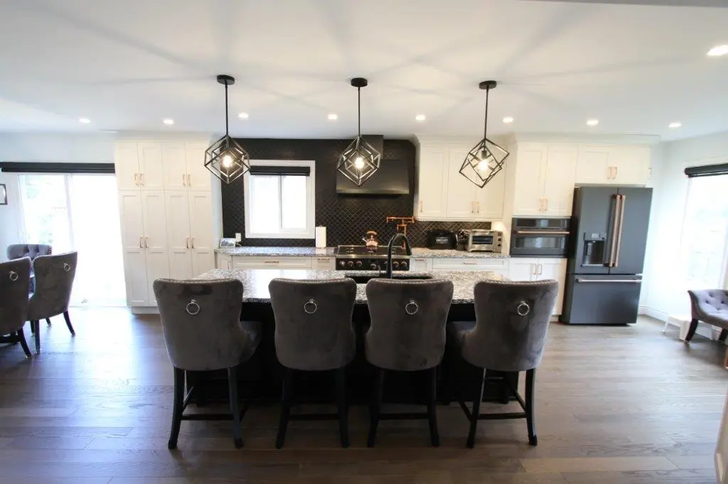 A modern kitchen with a large island featuring gray upholstered chairs, pendant lights, white cabinetry, and stainless steel appliances.