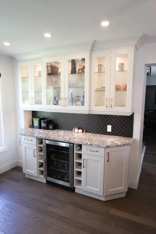 A modern kitchen with white cabinetry featuring glass doors on the upper cabinets, a quartz countertop, and a small wine fridge in the center of the lower section. The backsplash is a geometric pattern, and there are recessed ceiling lights enhancing the setting.