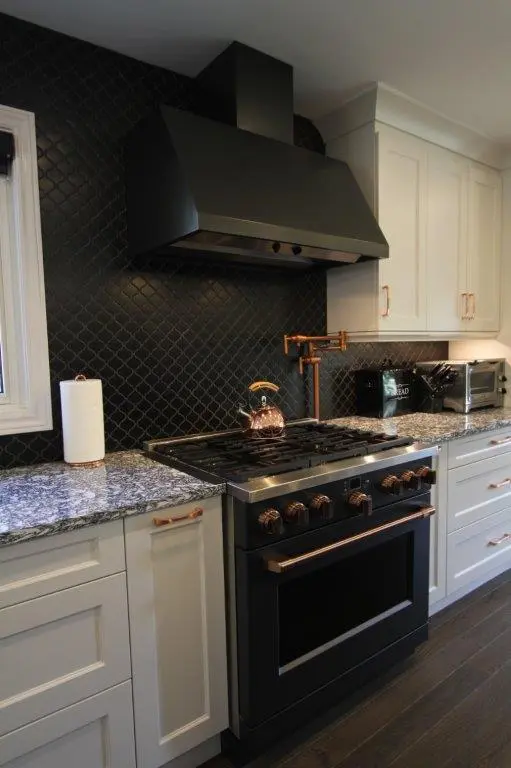 Modern kitchen with a black stove and hood, surrounded by white cabinets and a dark tiled backsplash. The countertop is speckled grey quartz, and copper accents are visible on the stove and faucet.