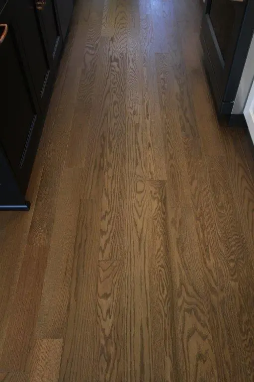 A close-up view of a wooden floor with a natural brown finish and visible grain patterns, bordered by dark kitchen cabinets on both sides.