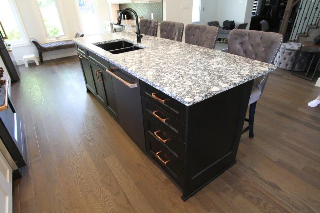 A modern kitchen island with a speckled quartz countertop, featuring an integrated sink and faucet, with dark cabinetry below. The island is surrounded by upholstered chairs and set on hardwood flooring.