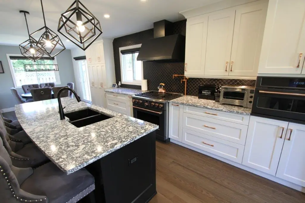 Modern kitchen with white cabinets, a speckled quartz countertop island, and pendant lighting. The kitchen features a black backsplash, a large oven with a range hood, and a dining area in the background.