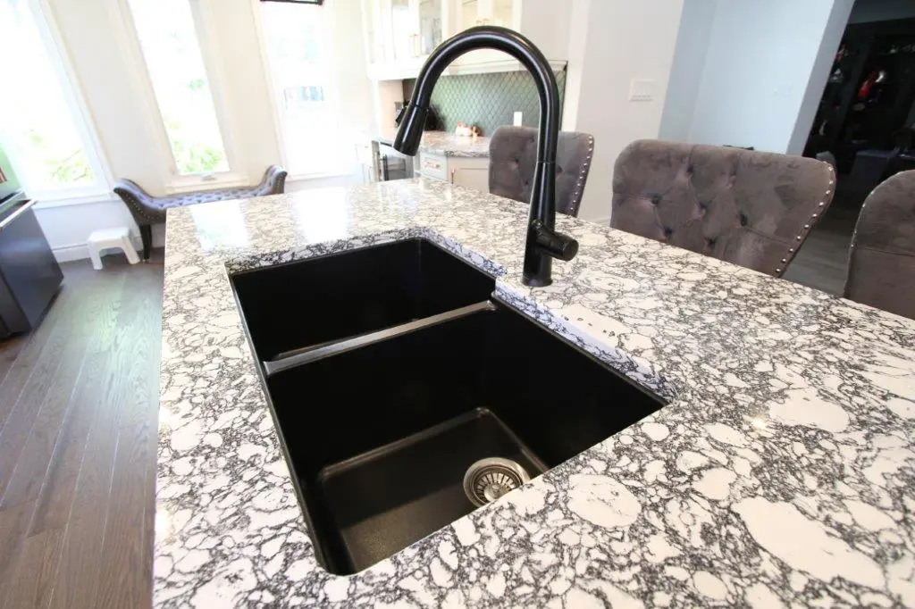 A modern kitchen sink with a black faucet is set into a quartz countertop with a distinctive black and white pattern. The faucet is tall and curved, and the sink is a deep, rectangular black basin. The surrounding kitchen area features gray cushioned chairs and large windows in the background, providing natural light.
