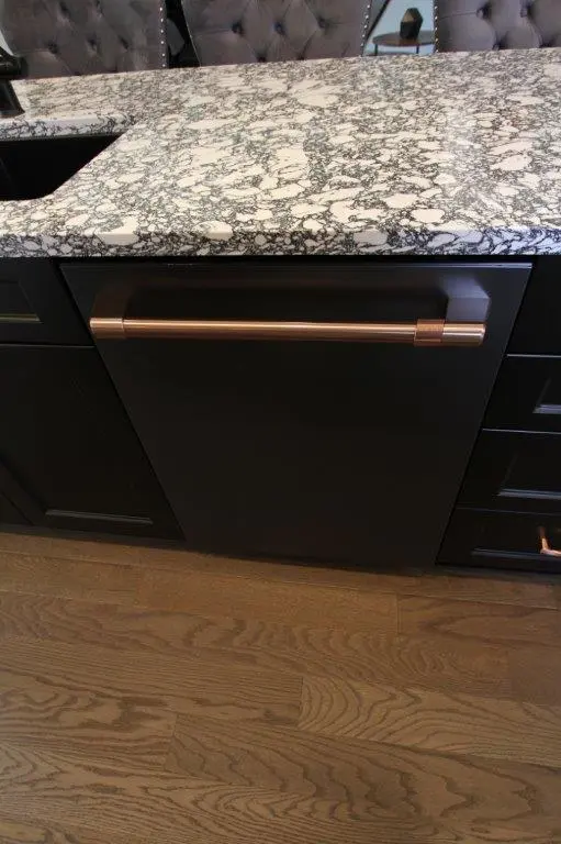 A kitchen area featuring black cabinetry with a marble-patterned countertop and a modern dishwasher with a copper handle, set on a wooden floor.