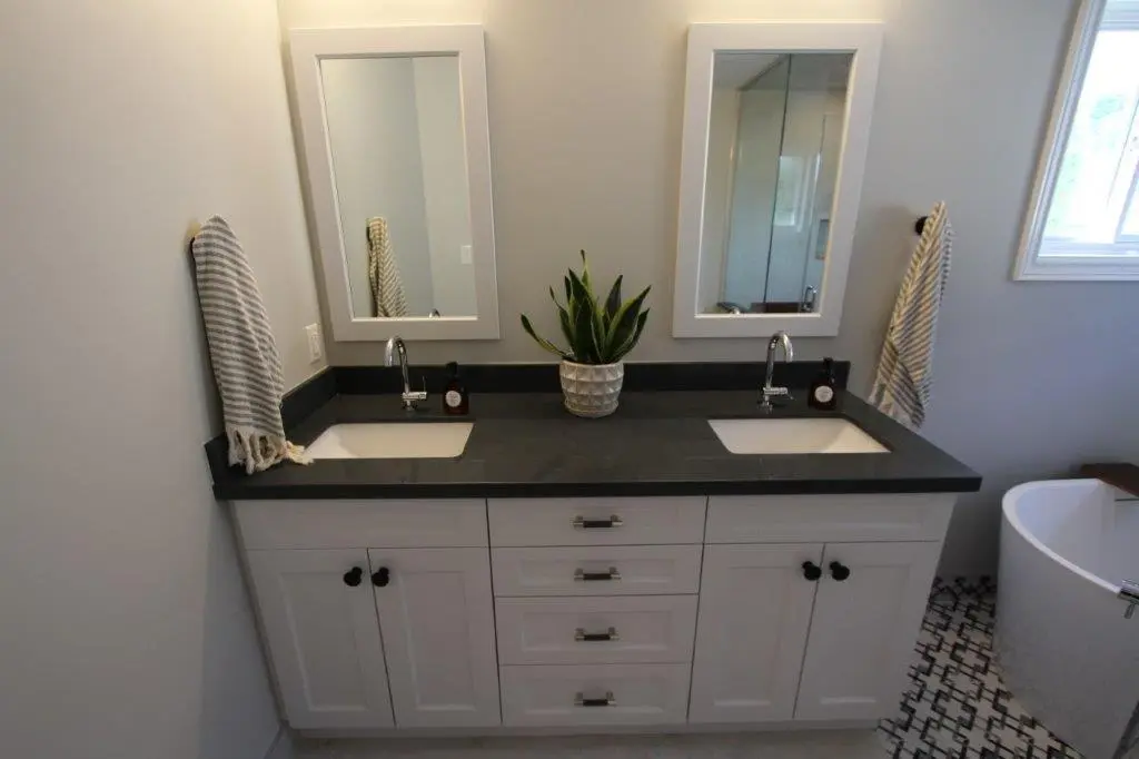 A modern bathroom with a double-sink vanity featuring dark countertops, dual mirrors, striped towels, a potted plant, and a visible portion of a bathtub.