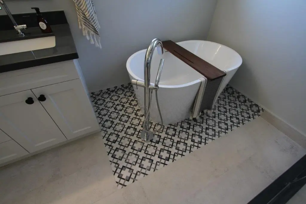 A modern bathroom featuring a white freestanding bathtub with a wooden tray on top, set on a patterned black and white tile section of the floor adjacent to a white vanity cabinet with a black countertop.