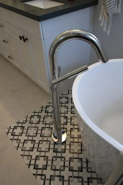 A modern bathroom with a white freestanding bathtub and a sleek chrome floor-mounted faucet. The floor features decorative black and white tiles, and a white vanity with dark countertop is visible in the background.