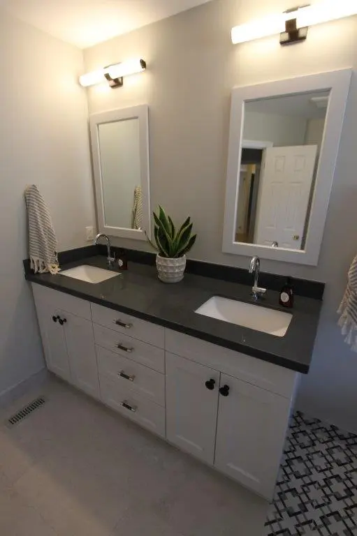 A modern bathroom vanity with a dark countertop featuring two sinks, each with its own mirror and light fixture above. Between the sinks is a small potted plant. The vanity has white cabinets and drawers, and the floor is partially tiled with a geometric black and white pattern.