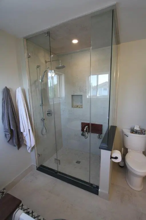 Modern bathroom with a glass-enclosed shower featuring a rainfall showerhead and handheld shower, adjacent to a toilet. Towels are hung on the wall, and there is a small recessed shelf in the shower.