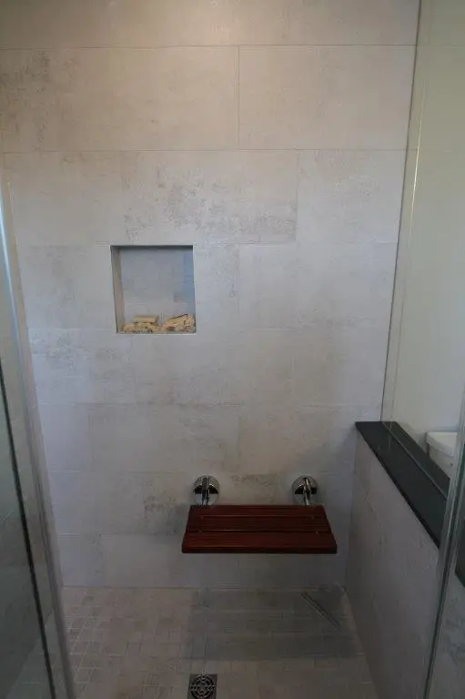 A modern shower with light gray tiles, featuring a recessed niche with soap, dual water controls, and a wooden foldable bench.