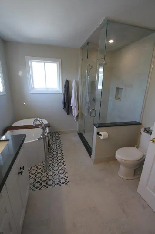 A modern bathroom featuring a white freestanding bathtub next to a glass-enclosed shower with a built-in niche. The floor has a patterned tile strip between the tub and the vanity. A window provides natural light, and towels hang on the wall.