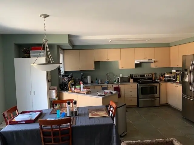 A before renovation kitchen and dining area with light wooden cabinets, stainless steel appliances, and a gray tiled floor. The dining table is covered with a gray tablecloth and surrounded by wooden chairs. A pendant light hangs over the table, and various kitchen items are placed on the countertops.